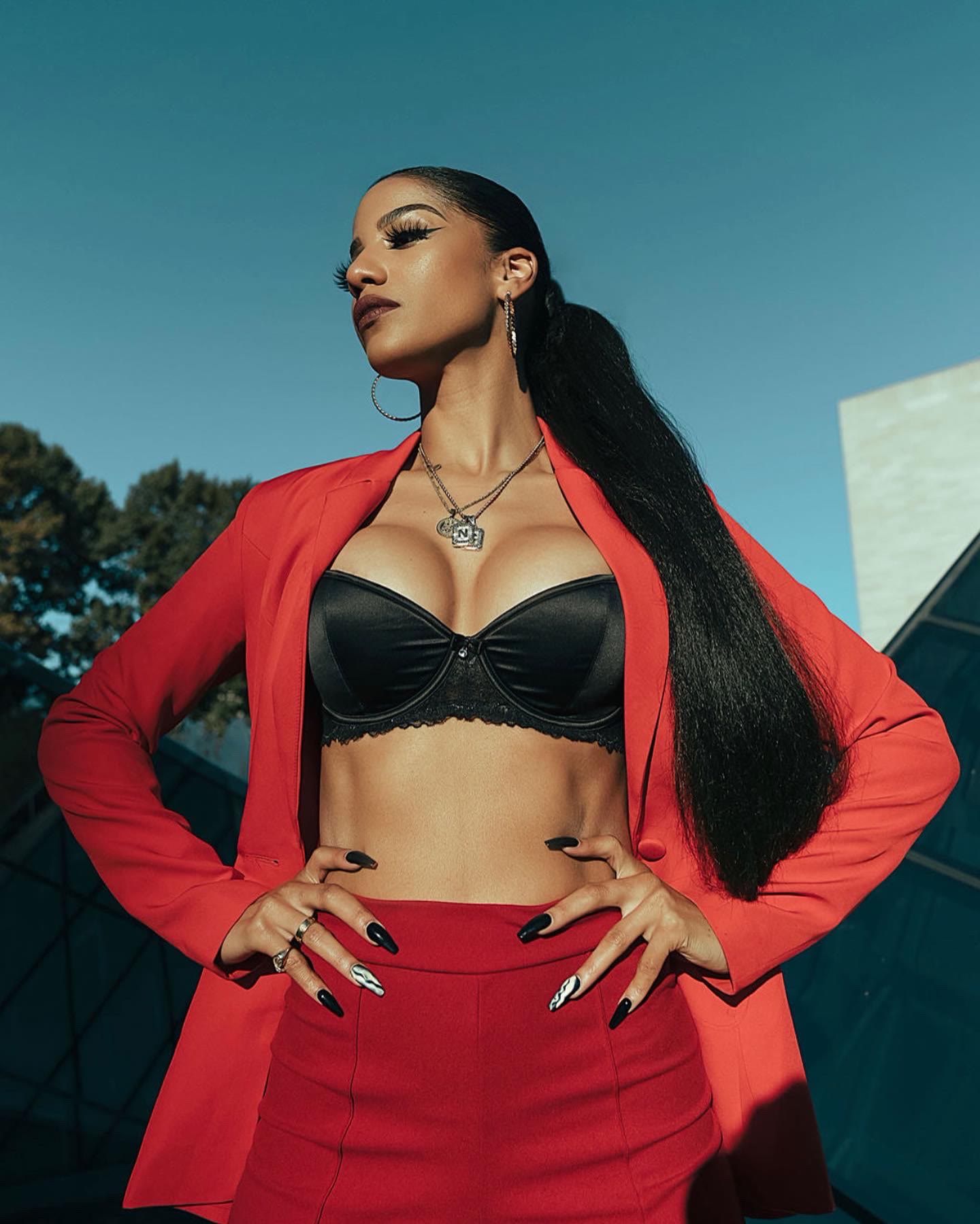 Low-angle shot of Christal in a red suit and black bra, hands on hips, long ponytail, blue sky
