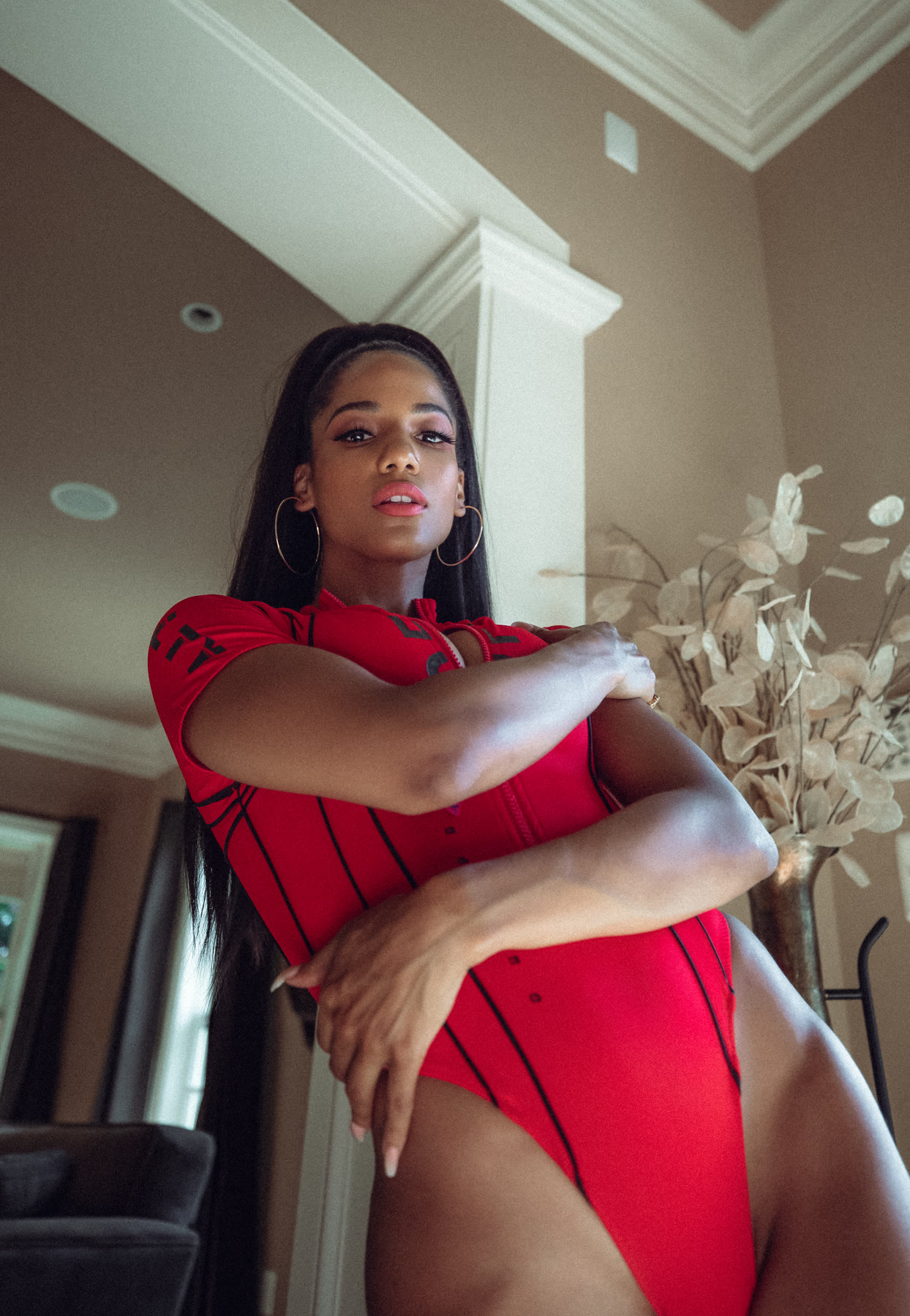 Low-angle shot of Christal in a red bodysuit with crossed arms and gold hoops
