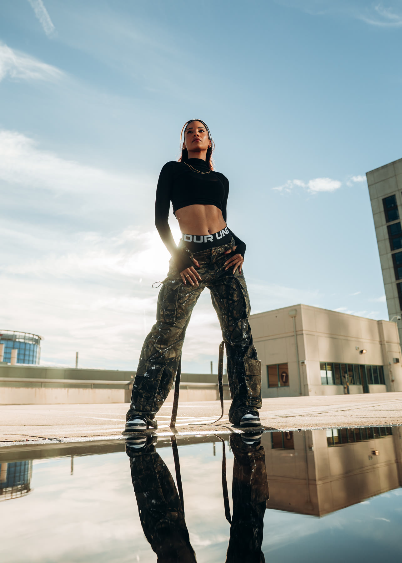 Low-angle shot of Christal on a rooftop in a black mesh crop top and camo cargo pants, reflected in a puddle