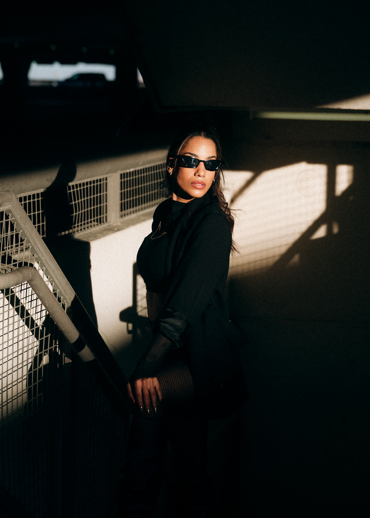 Christal in black sunglasses and a black blazer on a sunlit stairwell