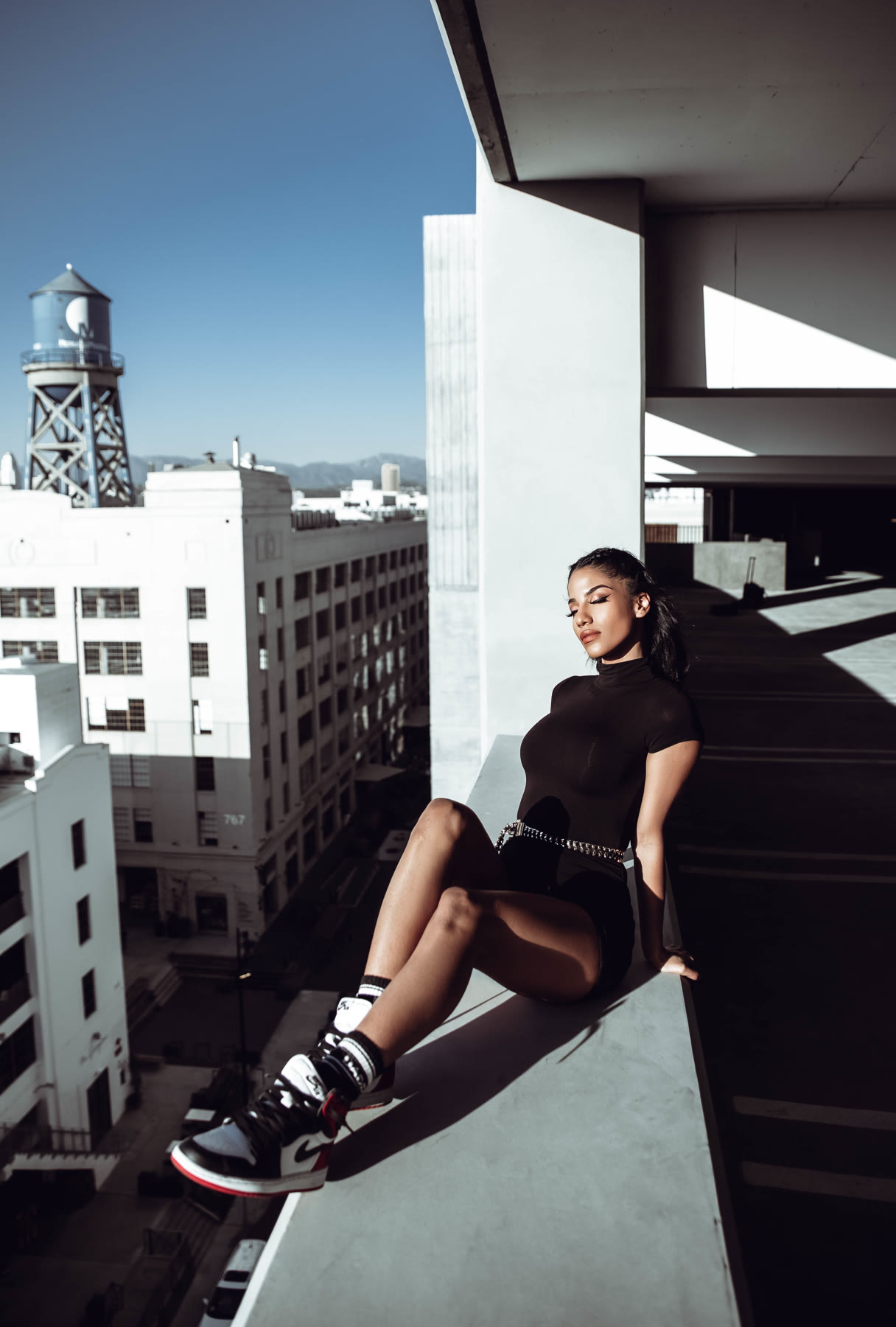 Christal sitting on a ledge high above the city in a black bodysuit and sneakers, eyes closed in the sun