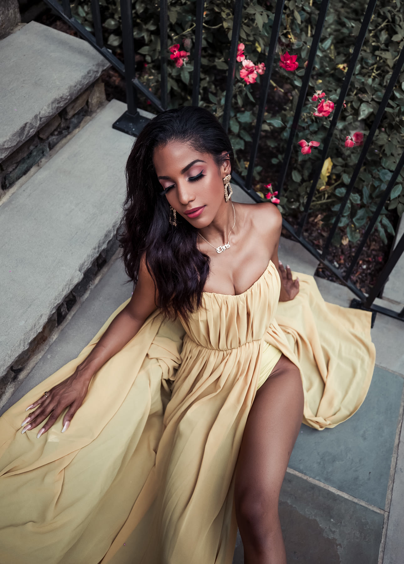 Christal seated on stone steps in the yellow gown, eyes closed, roses behind an iron railing