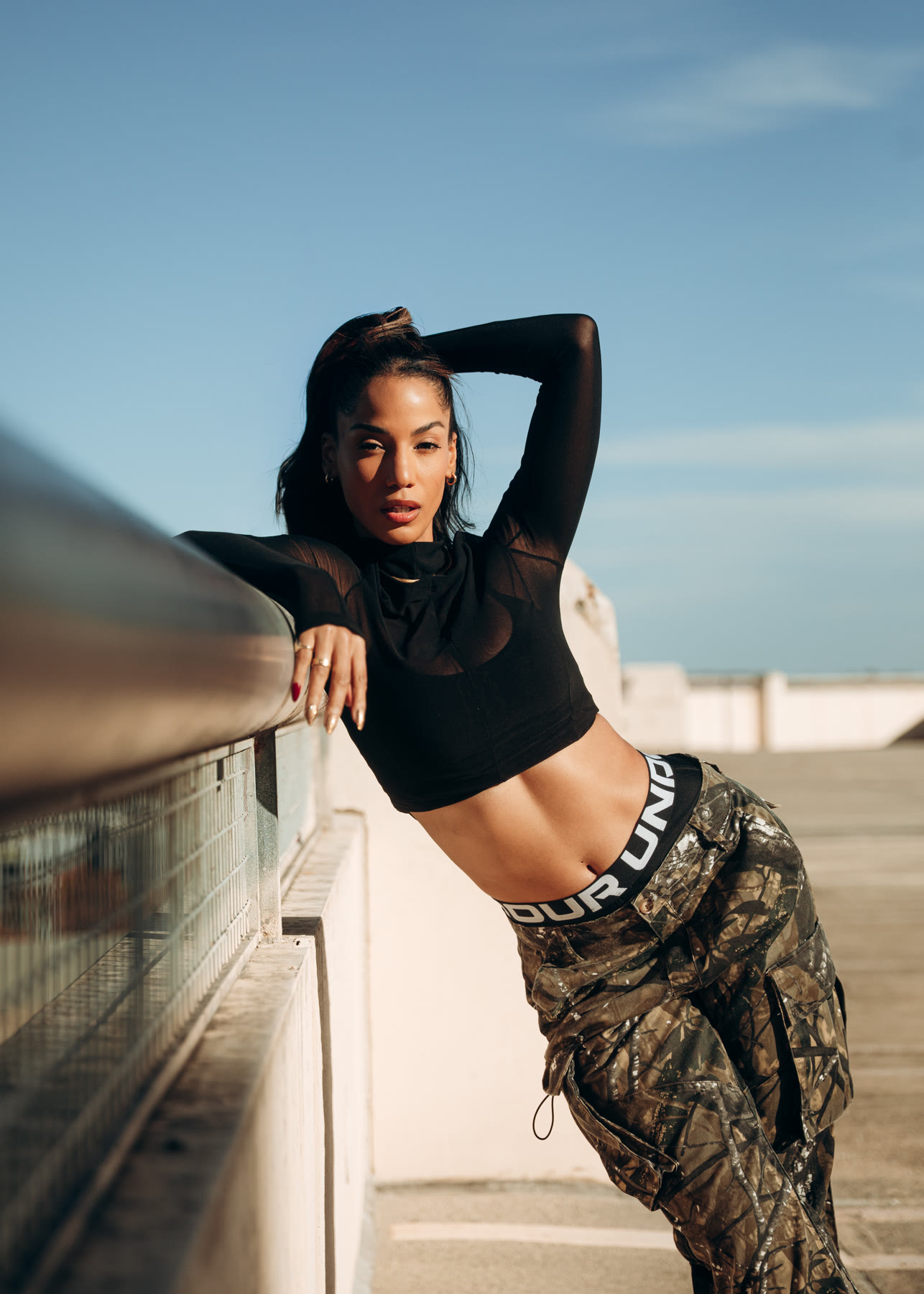 Christal leaning back against a rooftop railing, arm raised behind her head, blue sky behind