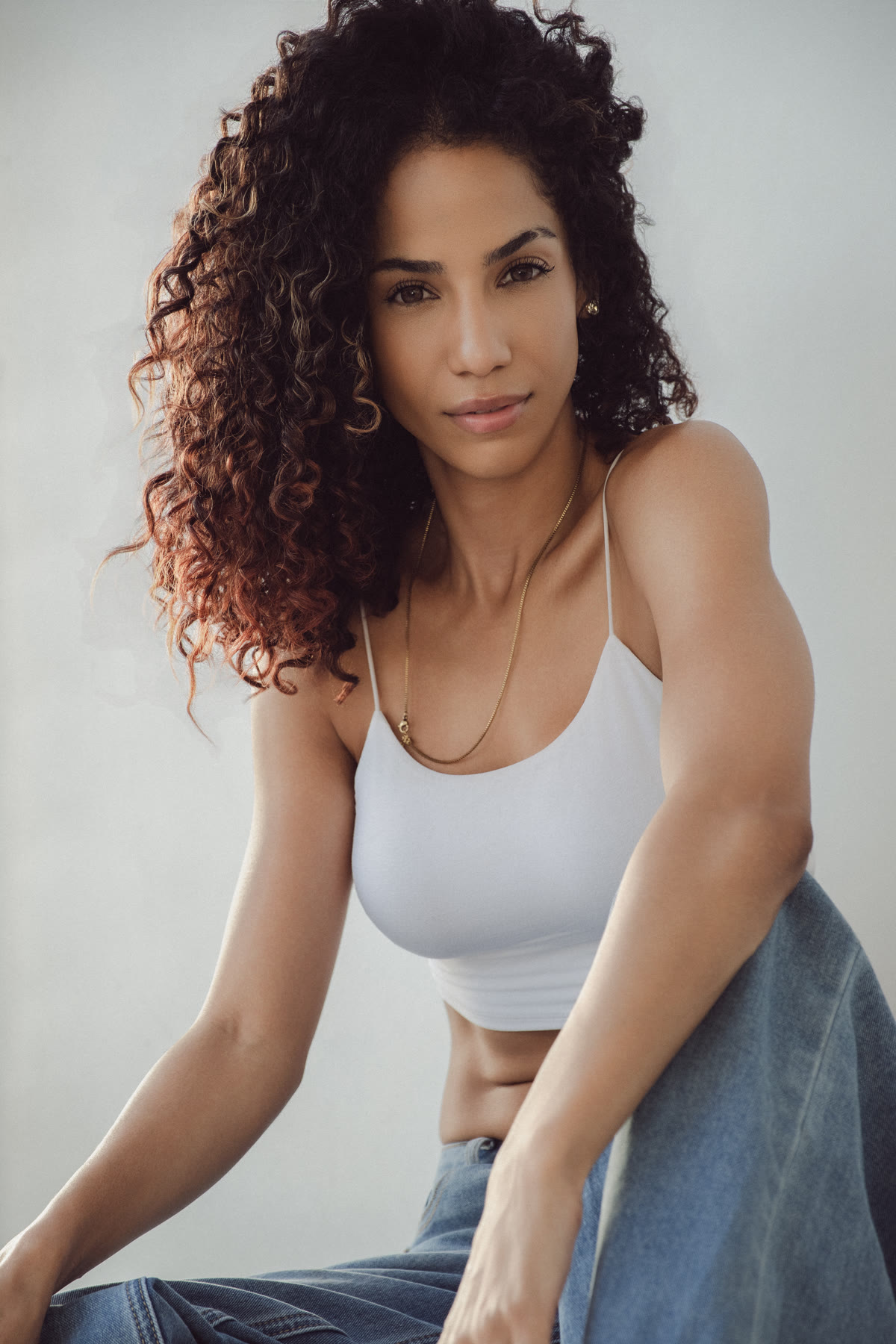 Christal seated, leaning forward toward the camera in a white crop top and jeans