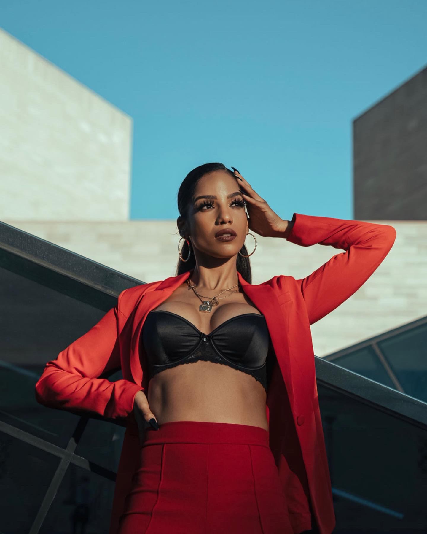Christal in a red suit, hand to her hair, against modern architecture and blue sky