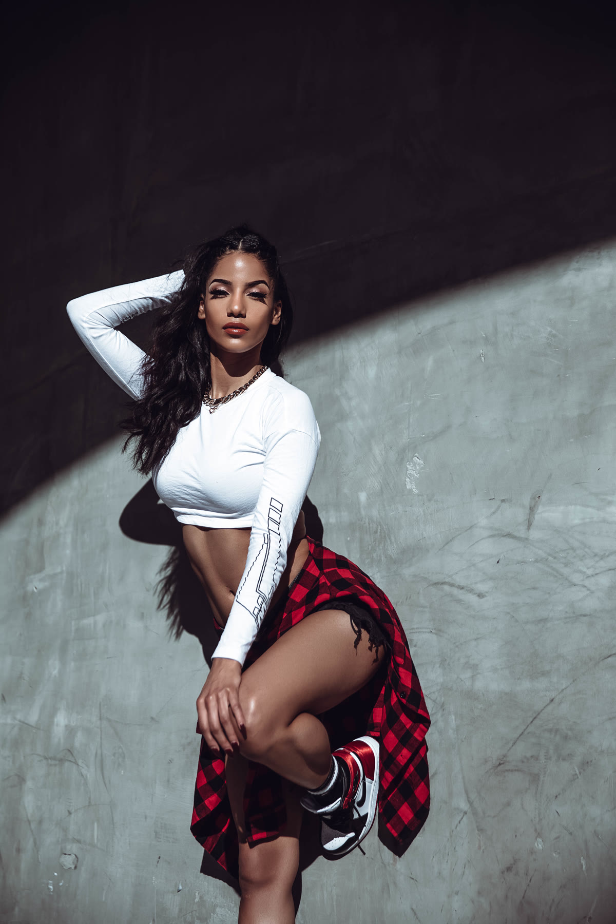 Christal against a concrete wall in a white crop top, plaid shirt at her waist and sneakers, knee raised