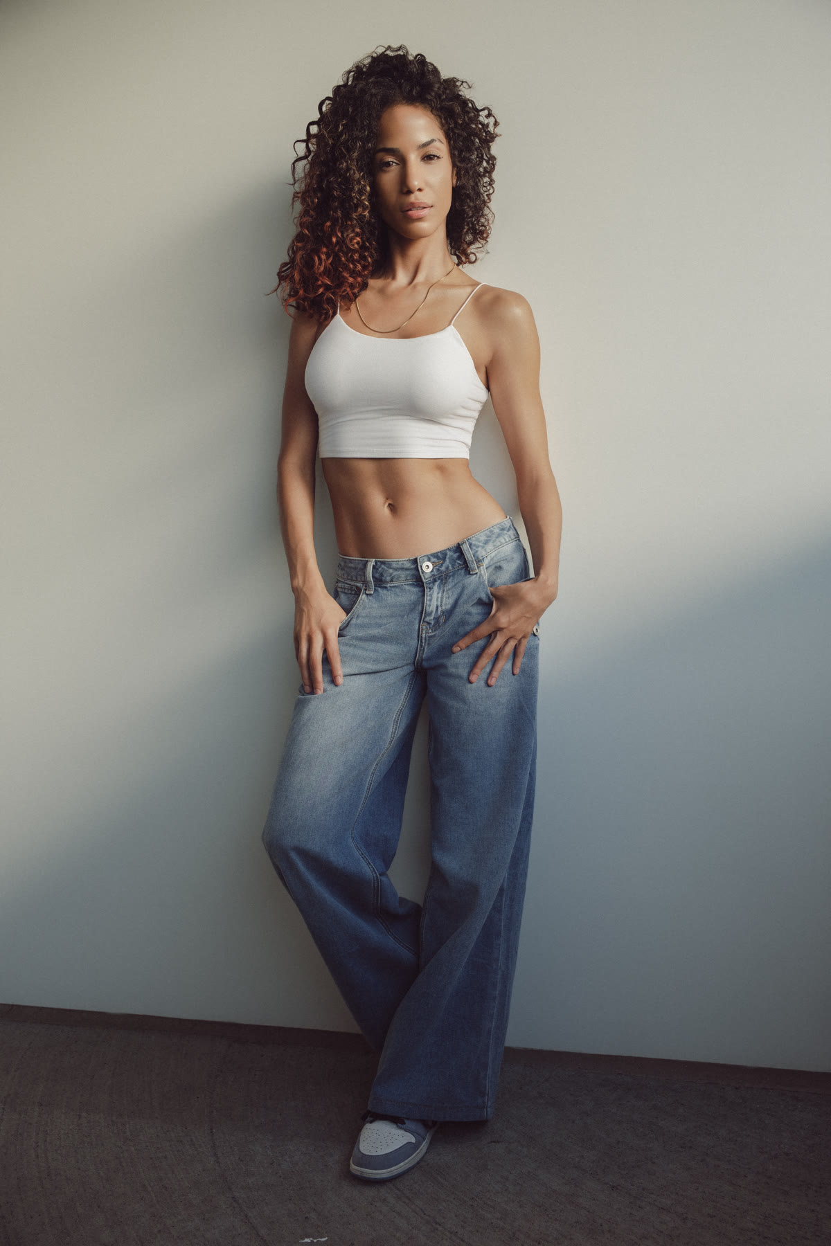 Full-length color shot of Christal leaning on a wall in a white crop top, wide-leg jeans and sneakers
