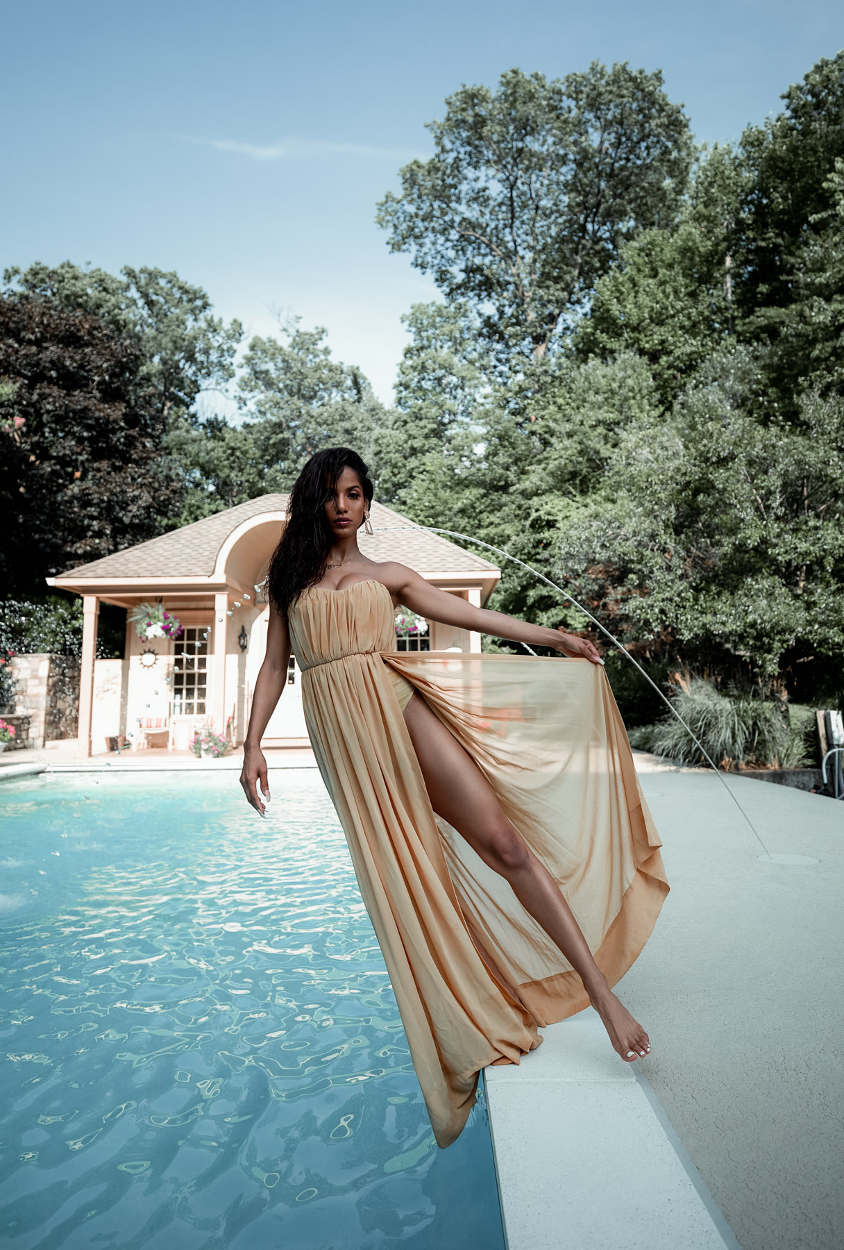 Christal stepping along a pool edge in a flowing strapless gold gown, holding the skirt out as a fountain arcs behind her