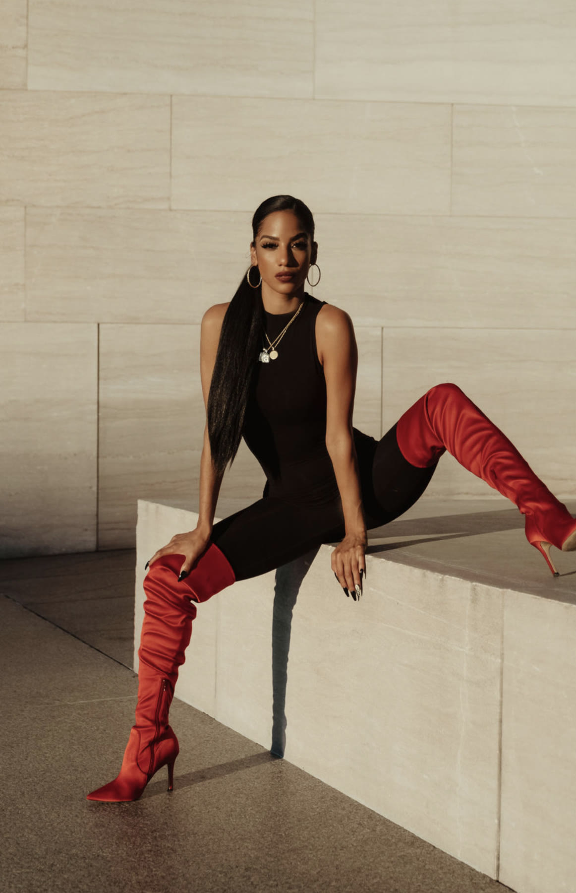 Christal seated on a stone ledge in a black bodysuit and red satin thigh-high boots