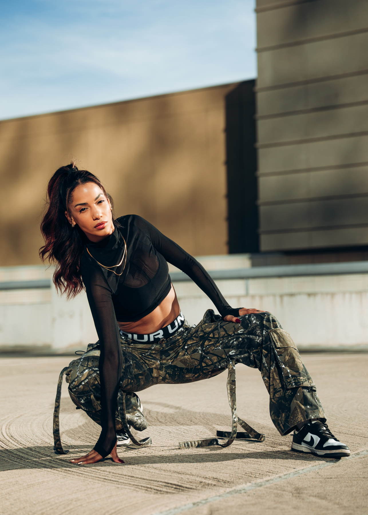 Christal crouching low in a black mesh top, camo cargos and black-and-white sneakers